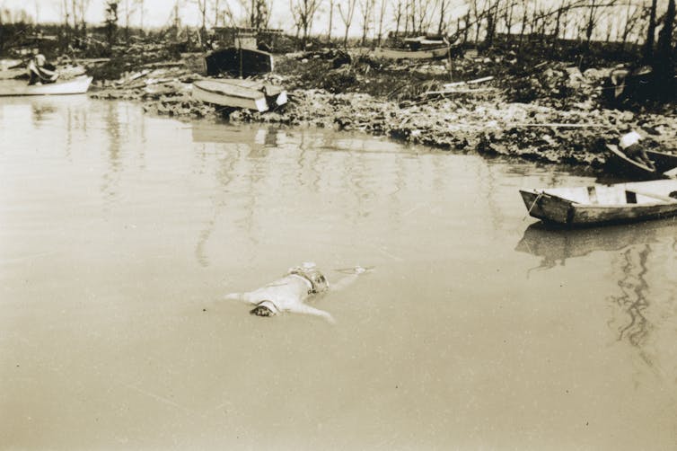 A dead man floats in brackish water with overturned boats and destroyed foliage on the shore in the background.