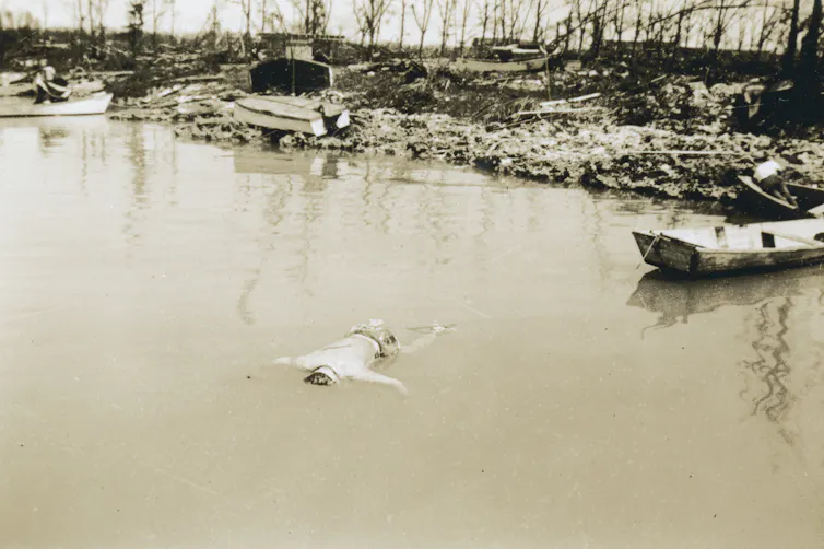 A dead man floats in brackish water with overturned boats and destroyed foliage on the shore in the background.