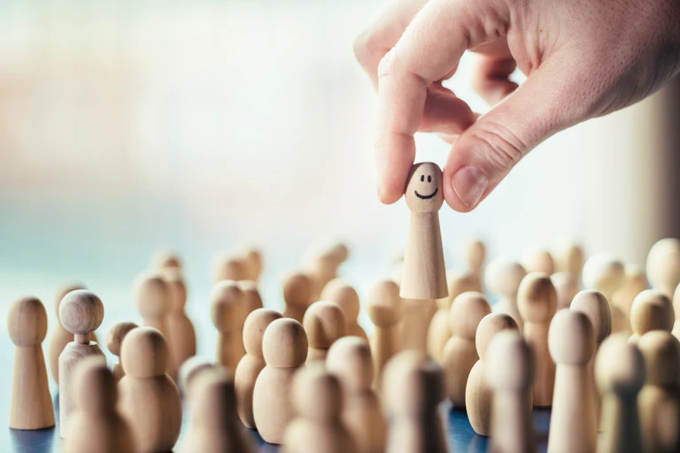 A hand picks up a wooden figure with a smiley face from among a group of other wooden figures.