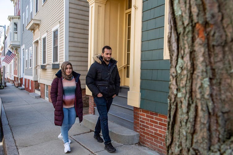 A woman and a man wear jackets and walk down a quiet street that has small buildings, including one with an American flag hanging off of it.