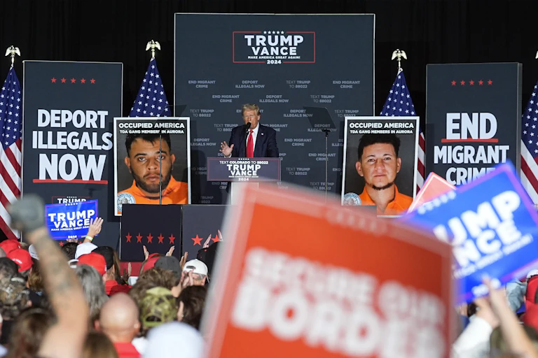 Trump speaks on stage in front of signs reading 'deport illegals now' and 'end migrant crime'