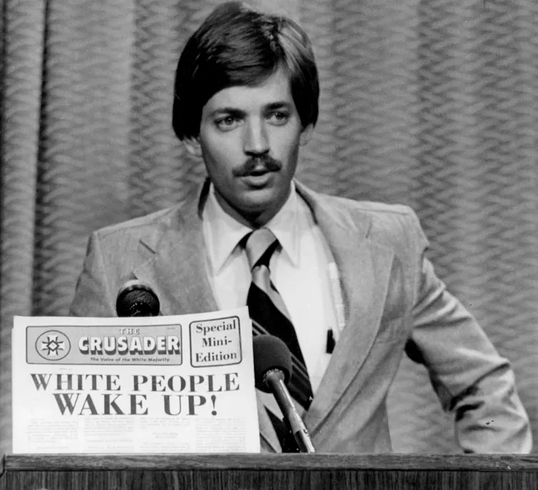 Man with a mustaff speaks at a microphone holding a newspaper reading, 'White people, wake up!'