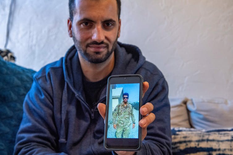 A man holds up a phone with a photo of himself wearing a camouflage uniform.