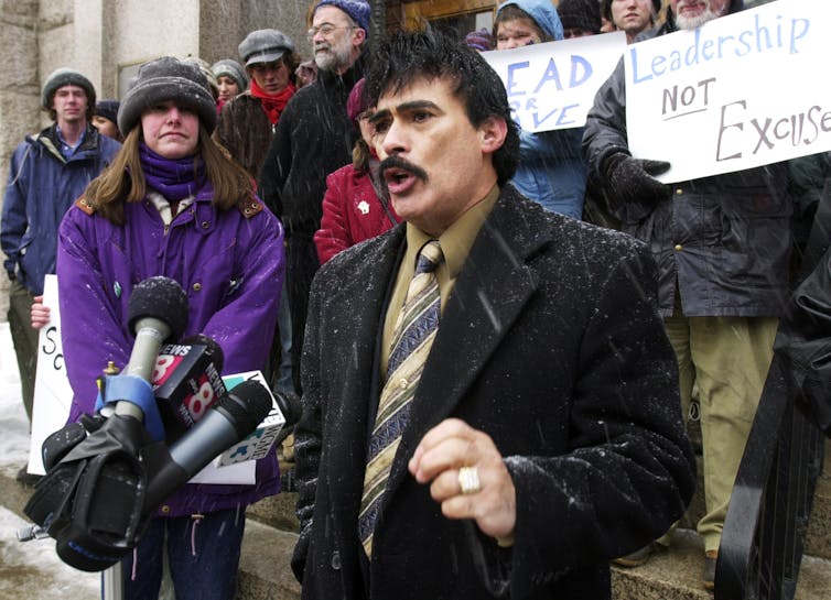 A man with a mustache speaks on the steps of city hall, surrounded by others; it is snowy.