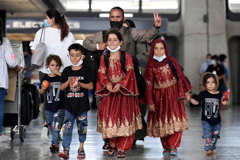 A man wearing a black face mask holds up two fingers and walks behind five children, including two girls who wear fancy looking maroon dresses.
