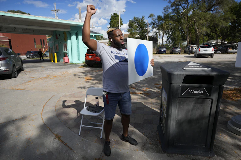 A man in a Kamala shirt holds a white sign with a blue dot on it.