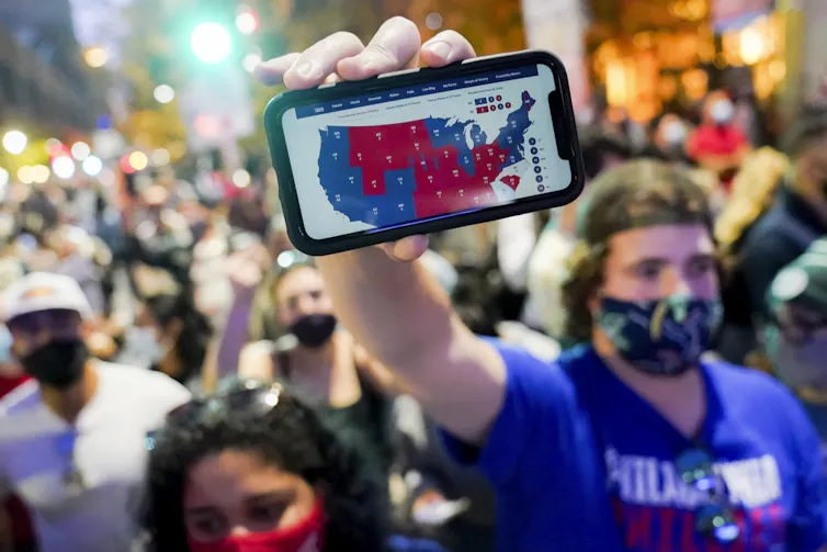A man in a Biden shirt holds up a map of the electoral college on his cell phone.