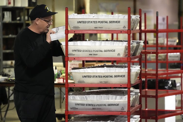 A man in a ballcap tends to several bins of mail-in ballots.