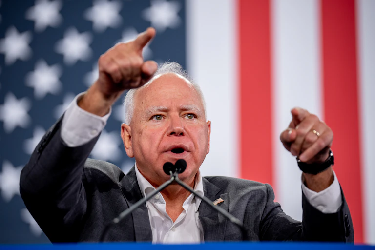 A man gestures at a lectern with two microphones while an American flag hangs vertically in the background.