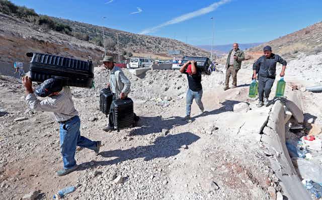Men carrying water bottles and luggage walk across rubble.