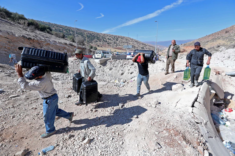 Men carrying water bottles and luggage walk across rubble.