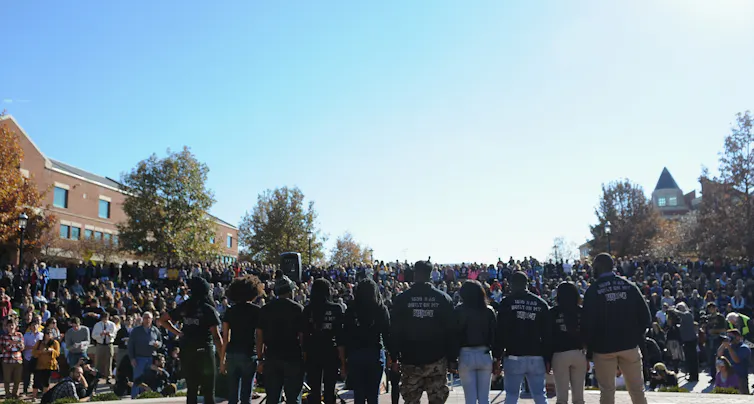 Black students stand holding hands on stage, their backs to the camera, facing a large crowd on a college campus