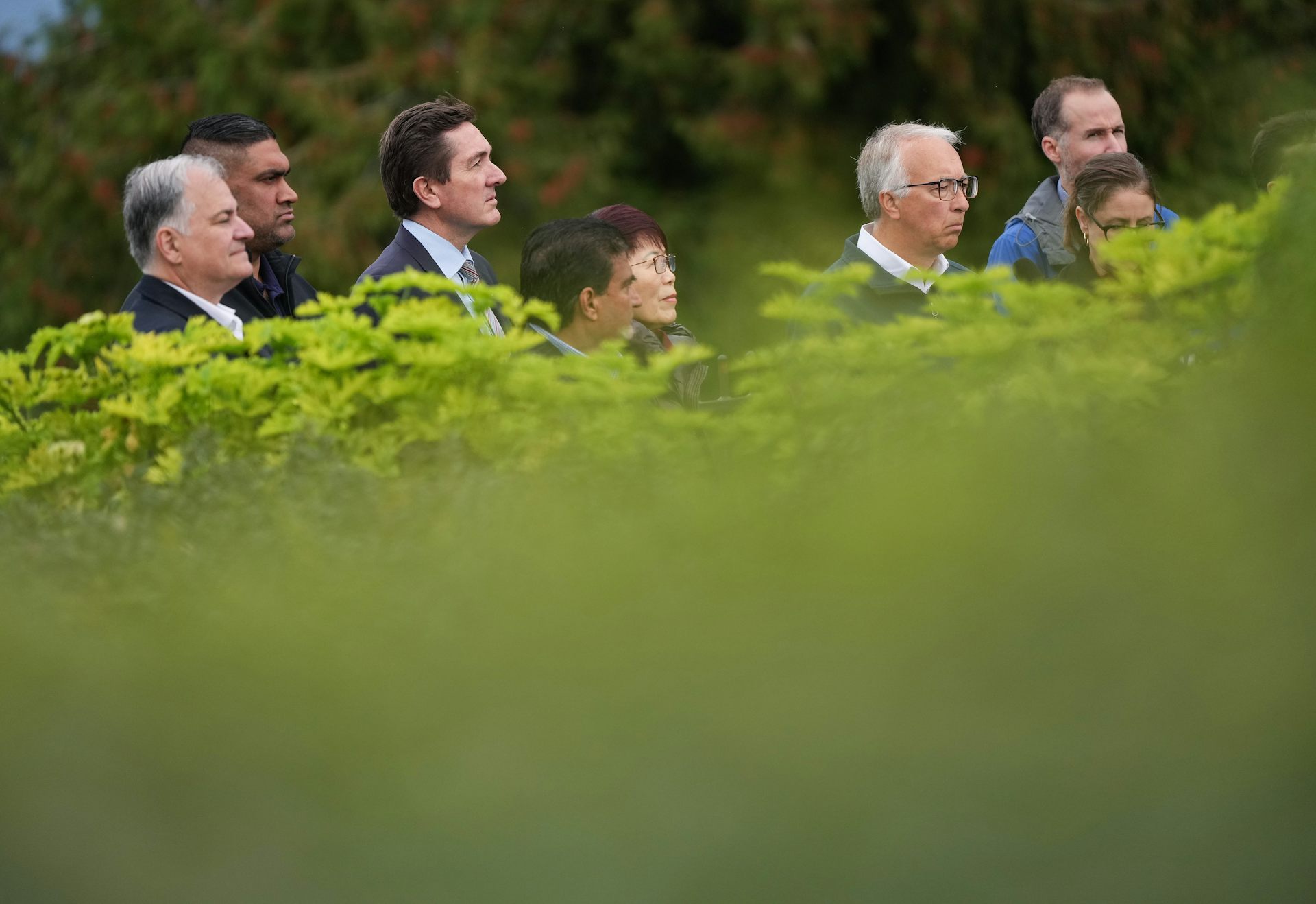 A group of politicians are photographed from behind a bright green hedge; only their heads and shoulders are visible.