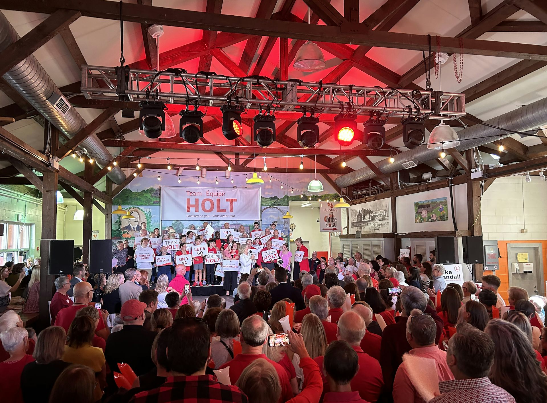 A room full of people with a red light illuminated above a stage, where a woman stands behind a podium.