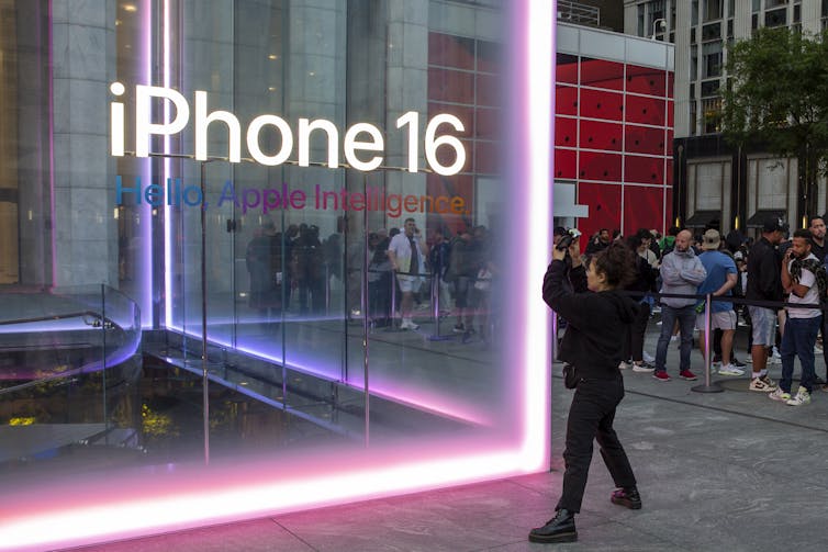 A person takes a photo of a giant glass cube with the words 'iPhone 16 Apple Intelligence' written across the front