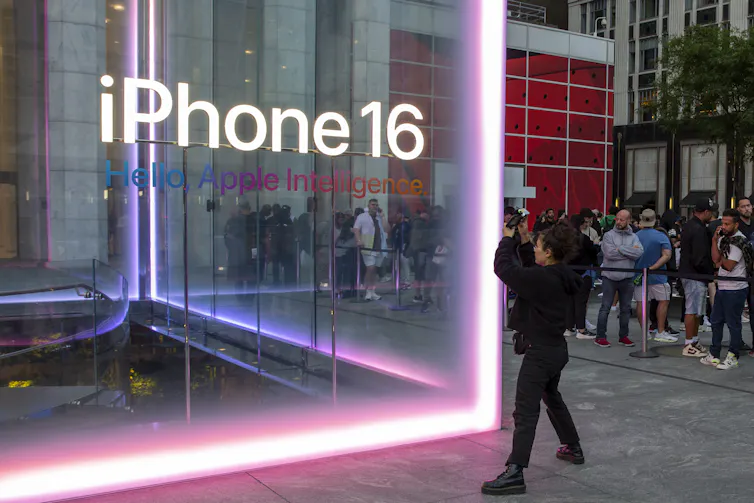 A person takes a photo of a giant glass cube with the words 'iPhone 16 Apple Intelligence' written across the front