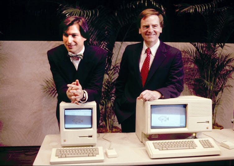 Two young men in suits stand behind an old-fashioned desktop computer