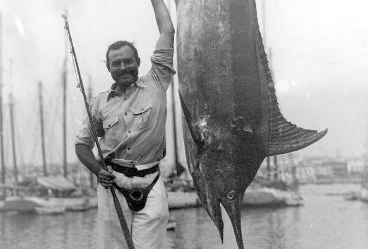 Mustached man holding fishing rod standing next to large fish.