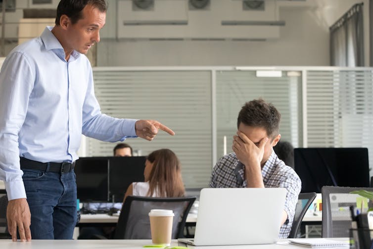 A man towers over a younger man sitting in front of an open laptop at a desk, pointing his index finger at him
