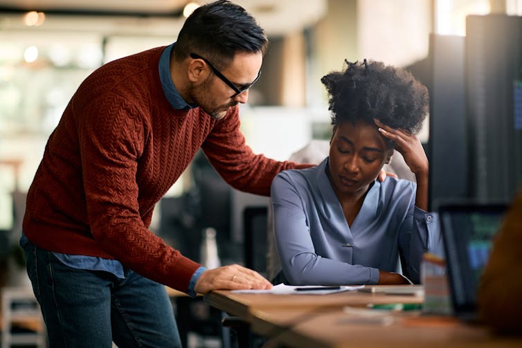 A man rests a hand on the shoulder of a woman who is sitting down at a desk with her head resting against her hand