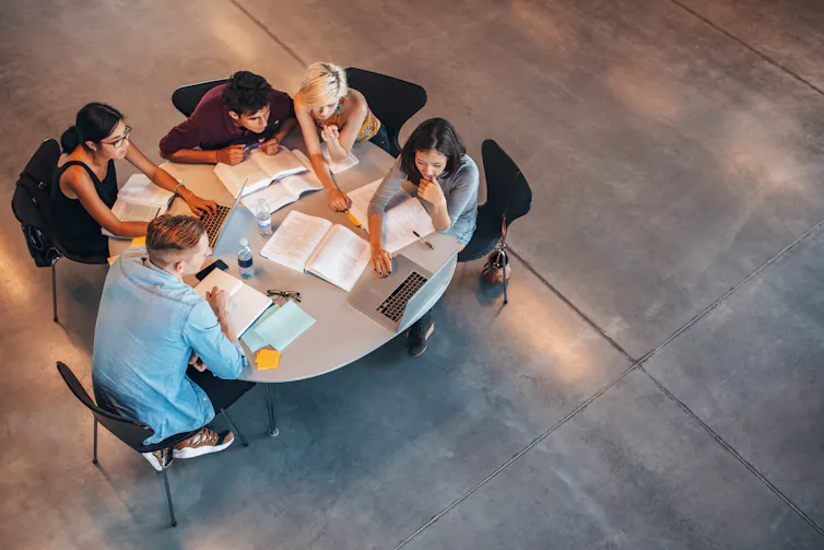 A group of young people sit around a table looking at a laptop. Books and papers are on the table.