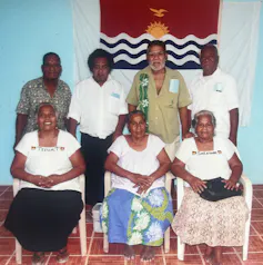 Seven members of the Kiritimati Association of Atomic Cancer Patients in front of the island's flag.
