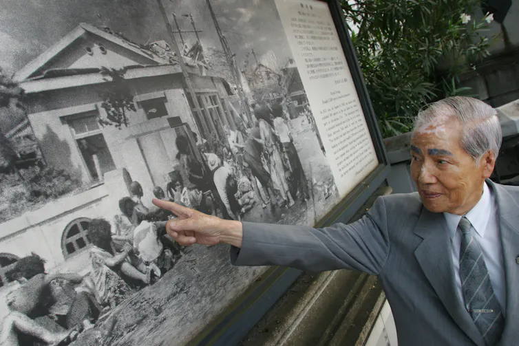 An old Japanese man in a grey suit who is a survivor of the Hiroshima bombing pointing to himself in photograph taken three hourse after the bombing.