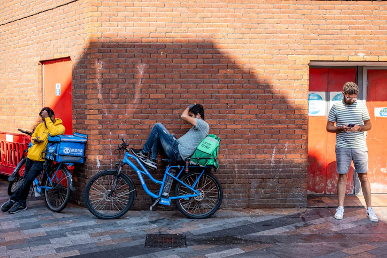 A man on his phone in an alleyway, next to two delivery cyclists taking a break on their phones.