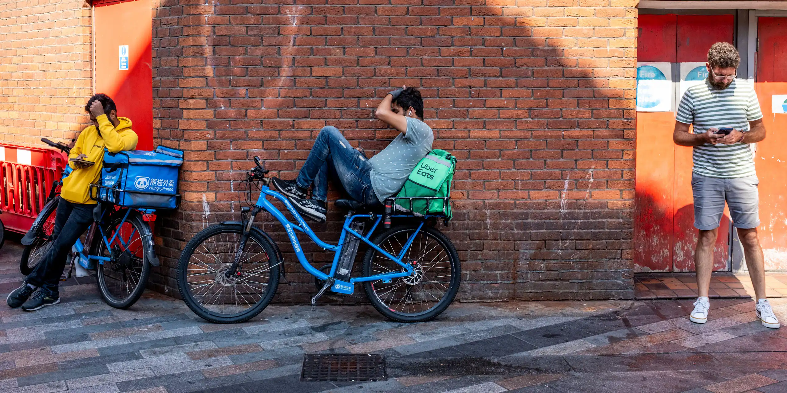 A man on his phone in an alleyway, next to two delivery cyclists taking a break on their phones.
