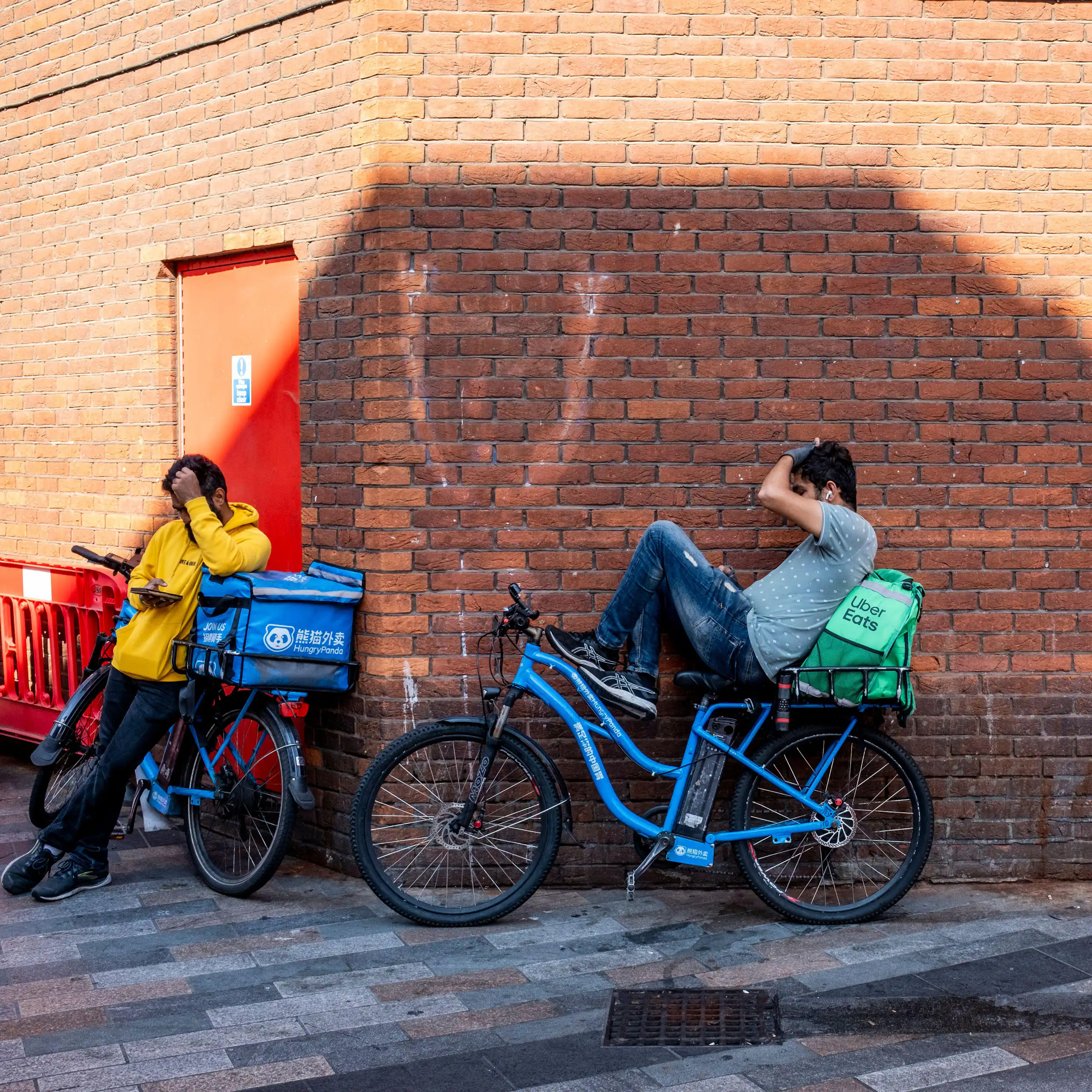 A man on his phone in an alleyway, next to two delivery cyclists taking a break on their phones.