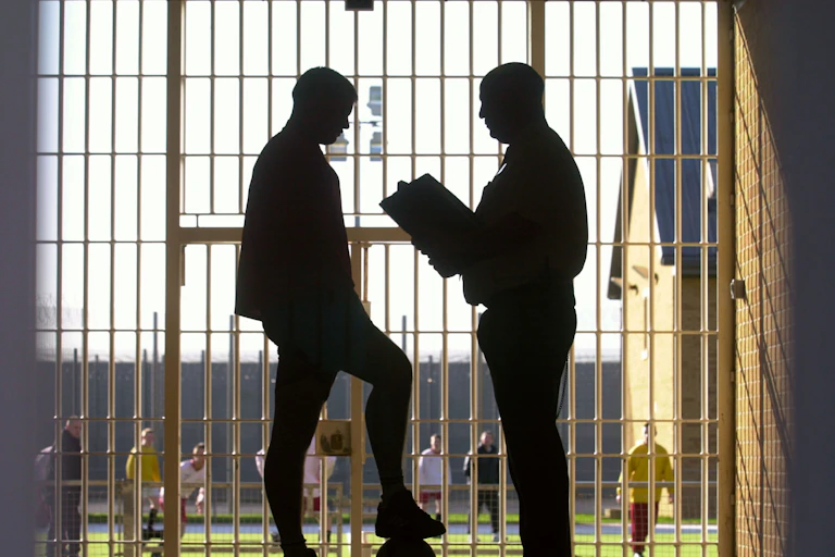 Silhouette of two men chatting, one is holding a clipboard and the other has his foot on a ball, there are prison bars and a football pitch behind them