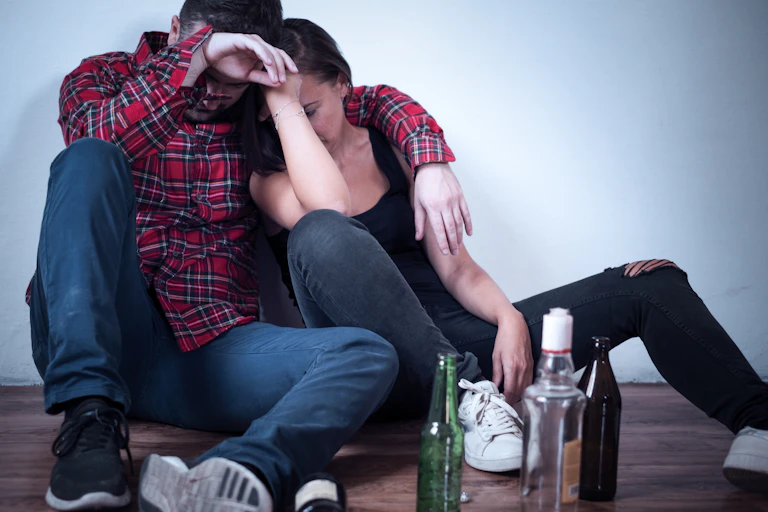 Two young people sit on the floor leaning against each other, with three empty alcohol bottles next to them.