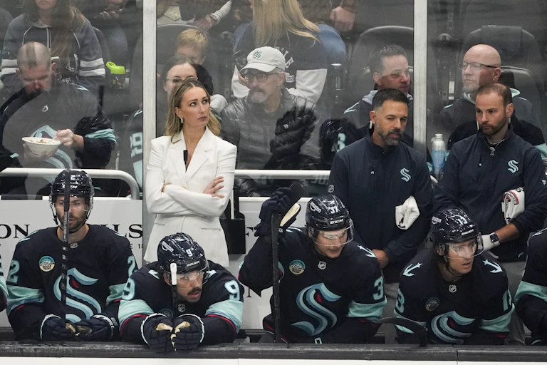 A white woman with blonde hair in a white suit stands behind a row of hockey players seated on a bench, with her arms crossed.