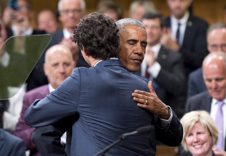 Two men, one a Black man, hug in the House of Commons and people look on smiling.