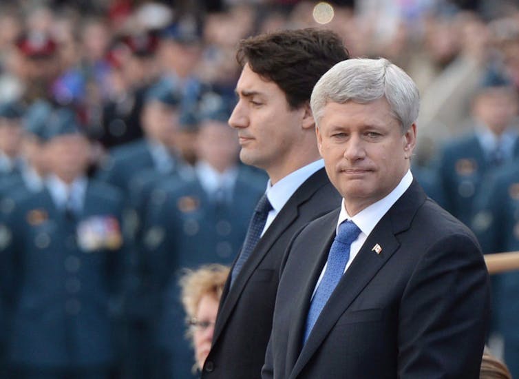 Two men, one with dark hair and the other older man with grey hair, prepare to place a wreath in front of them.
