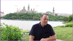 A man in a black shirt talks outdoors with the a river and Canada's Parliament buildings in the background.
