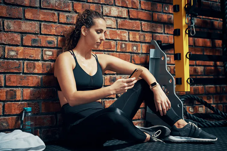 Woman sitting in gym wearing smart watch and looking at phone.