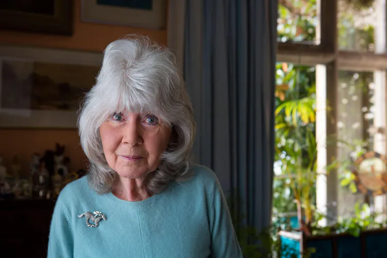 An elderly woman in a blue jumper in her living room with greenery at the window.