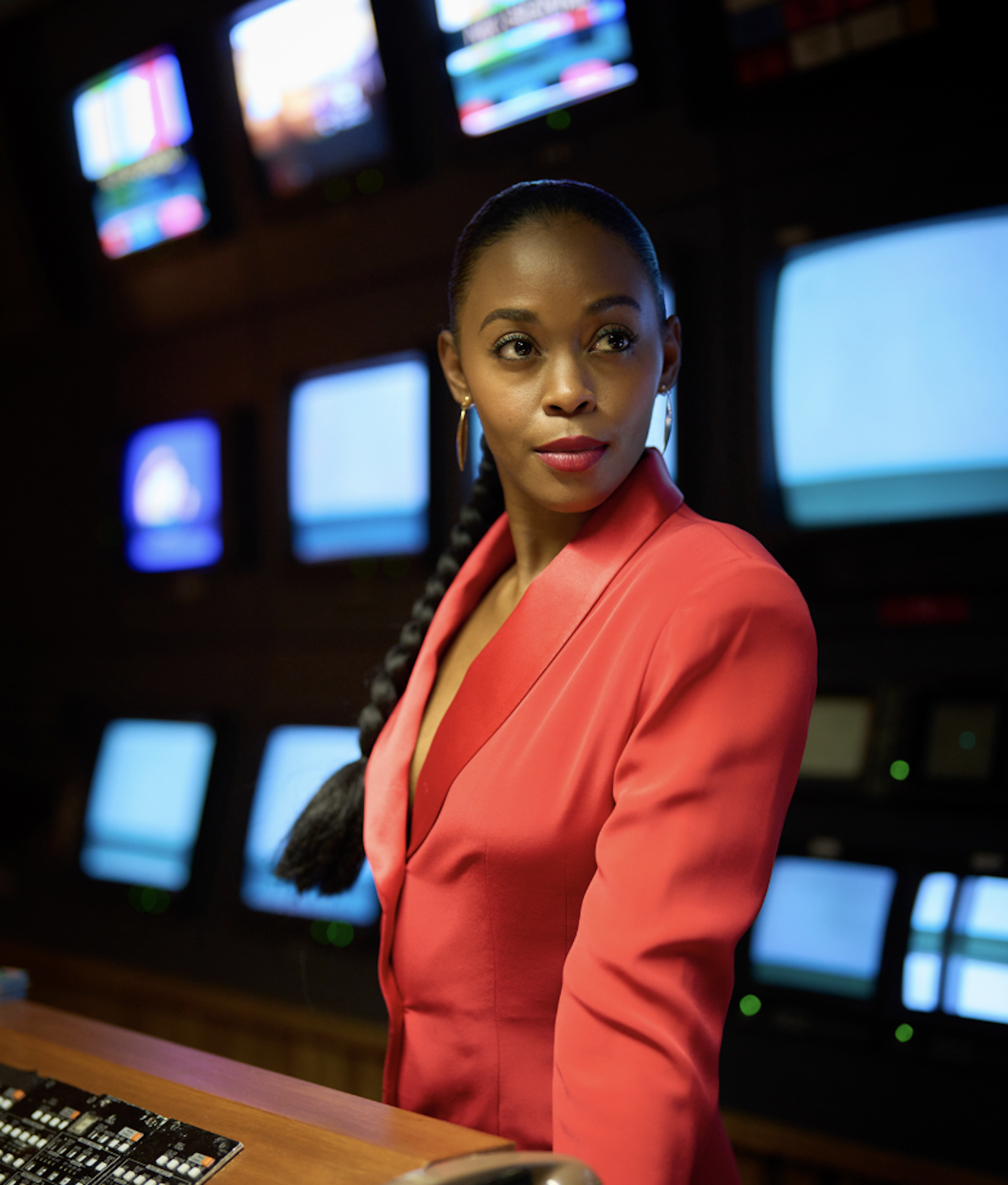 A young black woman dressed in a red suit in at a TV studio production desk.