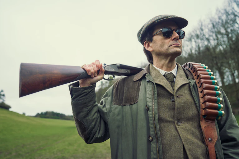 A man dressed in country sports attire with sunglasses holding a shotgun and ammunition over his shoulder.