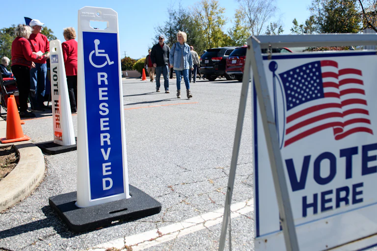 A large white sign with an American flag says 'Vote Hear' is positioned in a parking lot, as people walk toward it.