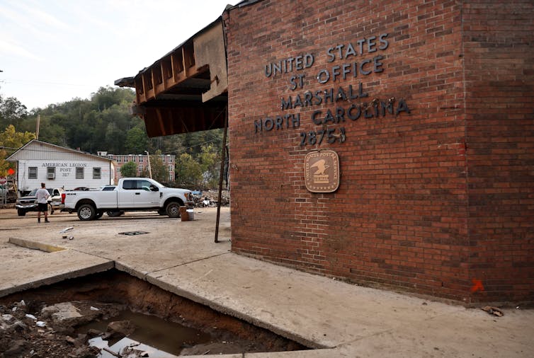 A brick building appears damaged on its front and in the ground next to it. The building has a sign that says 'United States Post Office' but some of the letters are missing.