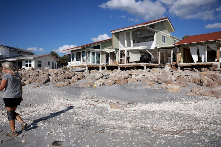 A woman walks on a gray path in front of several houses that appear empty and badly damaged.