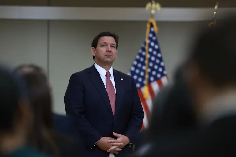 A white man in a blue suit and red tie looks pensive standing in front of an American flag.