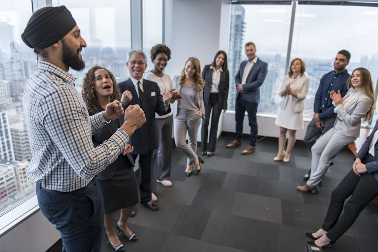 Around a dozen people in business clothing stand, smiling and chatting, in a room with big windows as a man with a turban speaks.