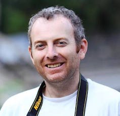 A smiling man in a white T-shirt with a camera strap around his neck.