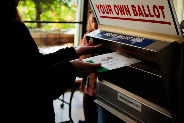 A person inserting a ballot into a box with a sign on it that says, in part, 'YOUR OWN BALLOT' AND 'INSERT BALLOT HERE.'