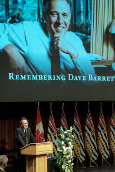 A photo of smiling man is seen on a large screen as a man delivers remarks at his funeral.