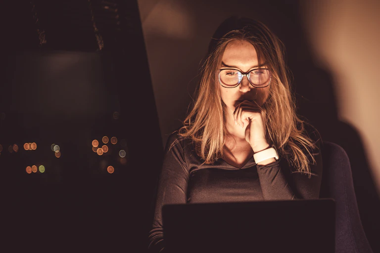 Woman wearing glasses with her left hand raised to her mouth, looking at her laptop in warm light.