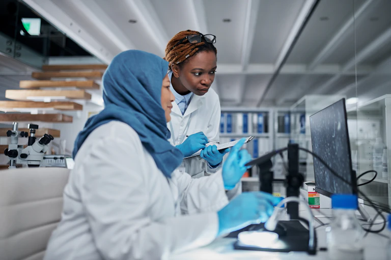 Two women in a lab wearing white coats look at a screen where they are developing a green energy innovation